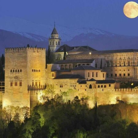 Vista de la Alhambra iluminada por la noche con la luna llena al fondo, destacando la majestuosidad del palacio en una visita nocturna