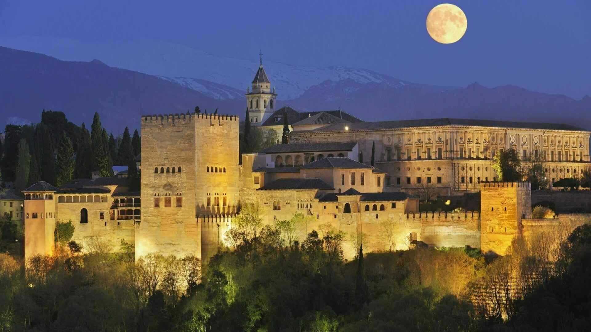 Vista de la Alhambra iluminada por la noche con la luna llena al fondo, destacando la majestuosidad del palacio en una visita nocturna